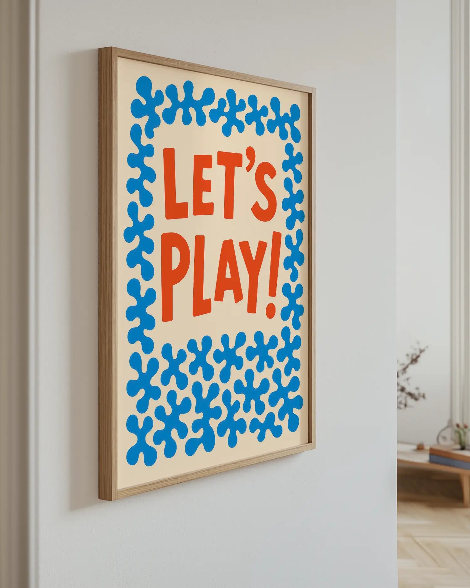 Inramad barntavla med texten "LET’S PLAY!" och blå dekorativa former, hängande på väggen i ett ljust barnrum. En glad och inbjudande väggdekoration.