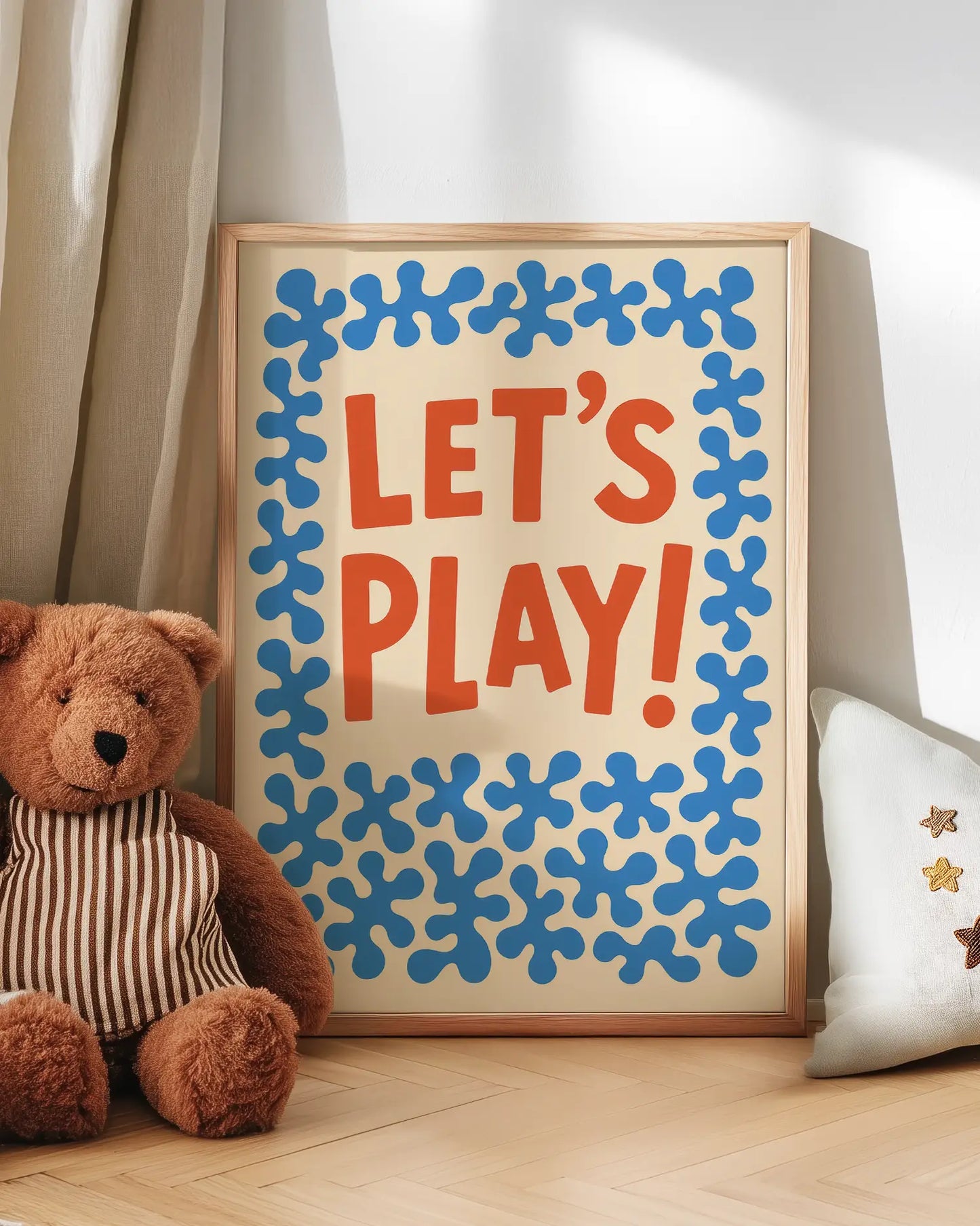 Inramad barntavla med "LET’S PLAY!" i orange och blå former, lutad mot väggen på golvet med en nalle bredvid. En mysig och lekfull miljöbild.