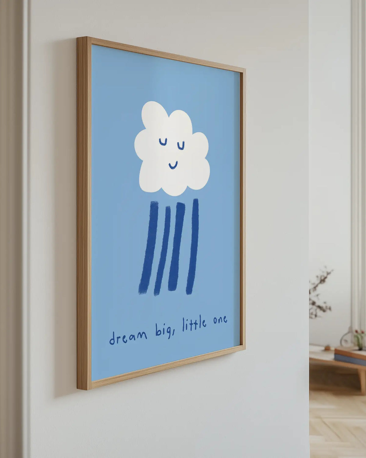 Barntavla med vitt moln med leende ansikte och texten “dream big, little one” på blå bakgrund upphängd på vägg med ekram. En charmig och drömsk väggdekoration.