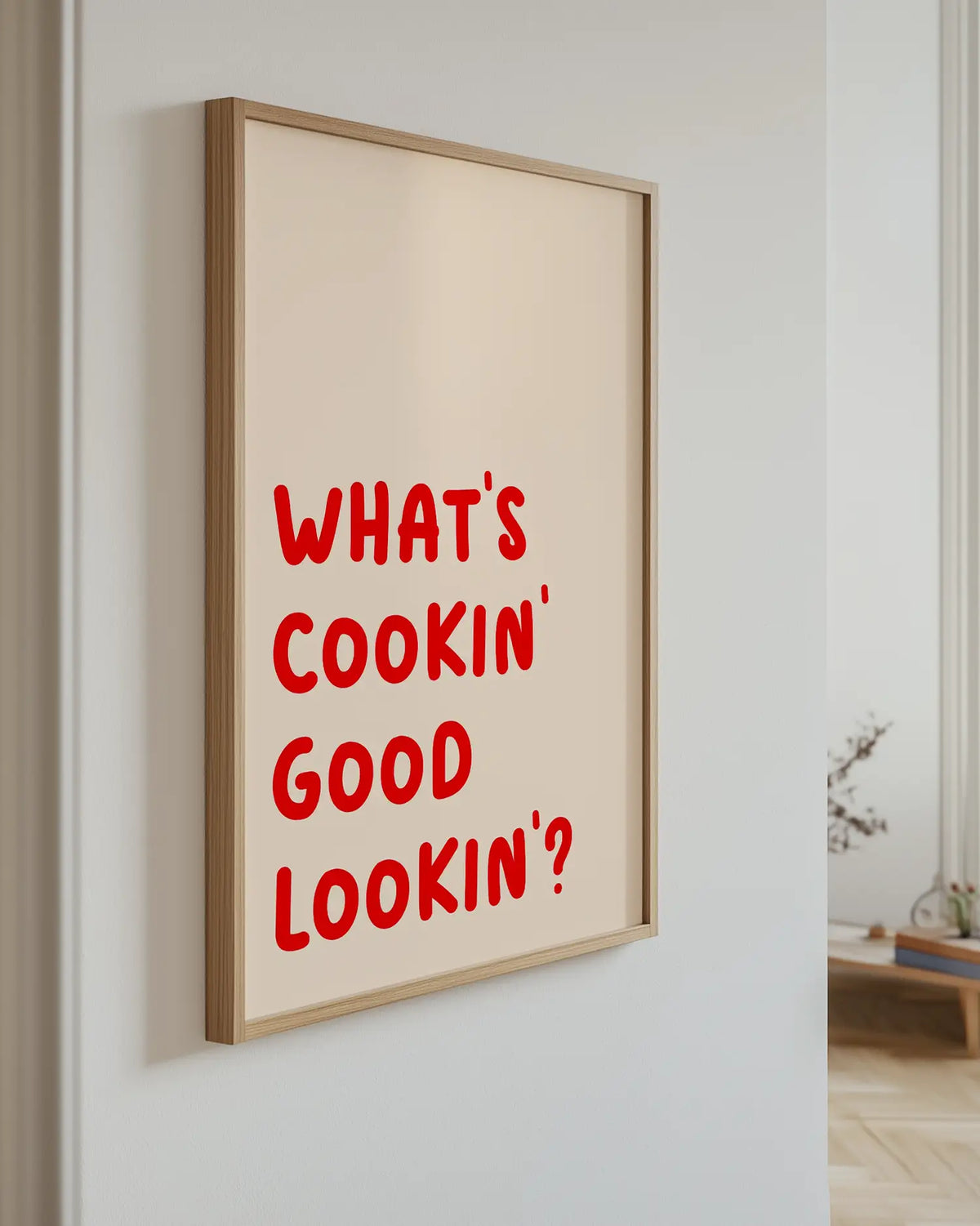 Barntavla med texten "WHAT’S COOKIN’ GOOD LOOKIN’?" i röd text mot beige bakgrund, upphängd på vägg med ekram. En elegant och modern presentation i ett ljust hem.