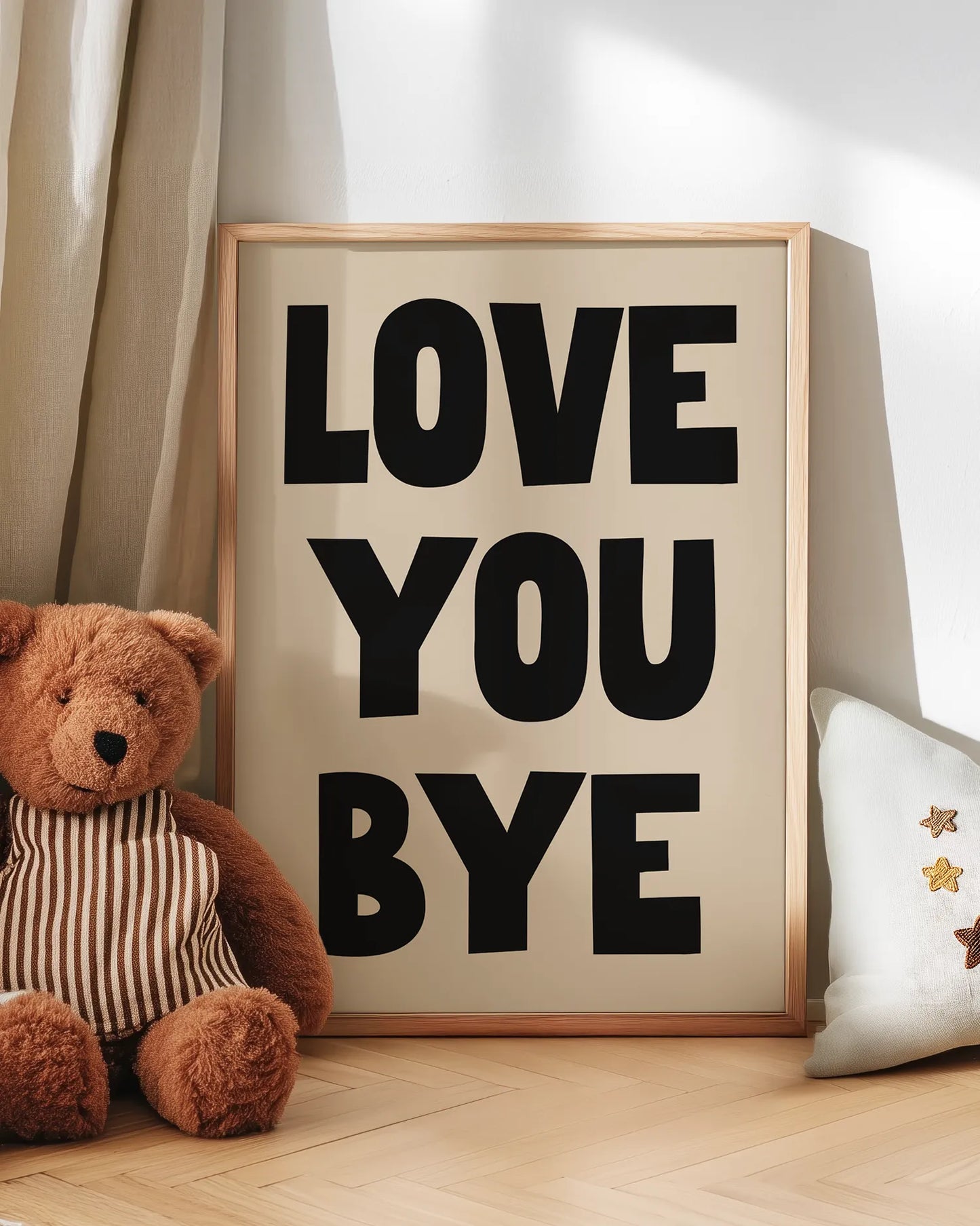 Inramad "Love You Bye"-poster i ekram stående på golvet bredvid en nalle och kudde i ett barnrum. Den beige bakgrunden och svarta texten skapar ett lekfullt och kärleksfullt uttryck, perfekt för barnrum eller familjehem.