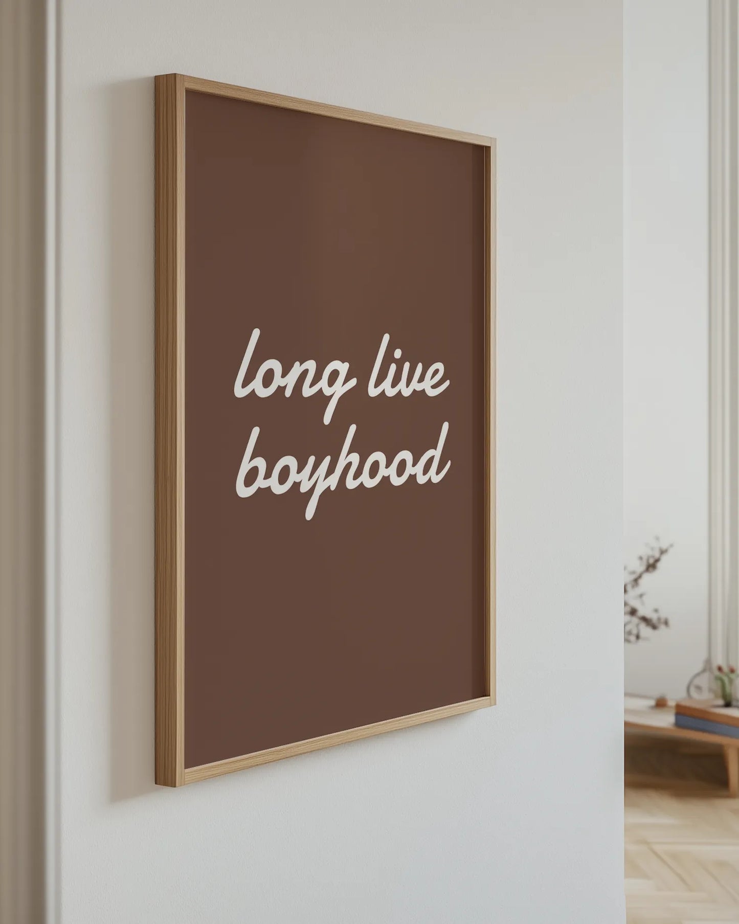"Long live boyhood"-poster i ekram hängande på en ljus vägg i ett modernt hem. Den bruna textpostern med vit handskriven stil skapar en varm och personlig känsla, perfekt som väggkonst i barnrum, lekrum eller vardagsrum med skandinavisk inredning.