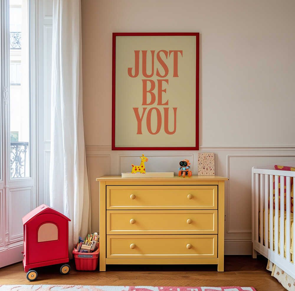 Inramad barnrumstavla med "JUST BE YOU" i orange text, upphängd ovanför en gul byrå i barnrum. En glad och inspirerande väggdekoration.