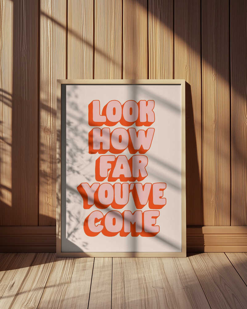 Inramad barnrumstavla med "LOOK HOW FAR YOU'VE COME" i rosa och orange, placerad på golvet framför träpanel. En varm och inspirerande inredningsdetalj.