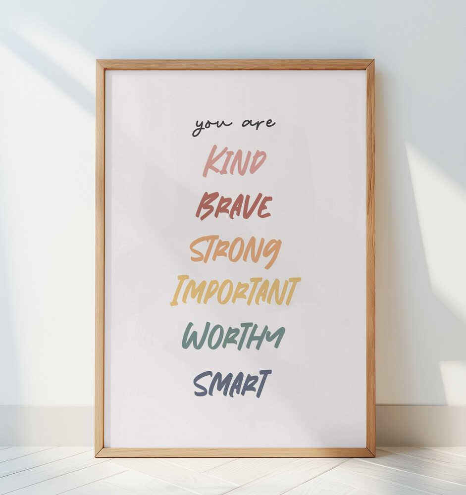 Barntavla med uppmuntrande budskap och texten "you are KIND BRAVE STRONG IMPORTANT WORTHY SMART" i ram, lutad mot en vit vägg. En stilren och trygg detalj i barnrummet.